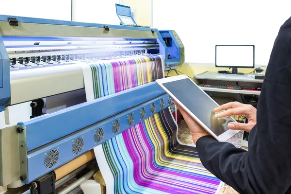 Foto de imprenta elaborando impresiones sostenibles mientras una persona con tablet diseña un tabloide.