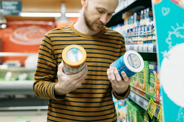 Hombre en pasillo de supermercado revisando y comparando diferentes tipos de etiquetas para productos en dos envases antes de decidir su compra
